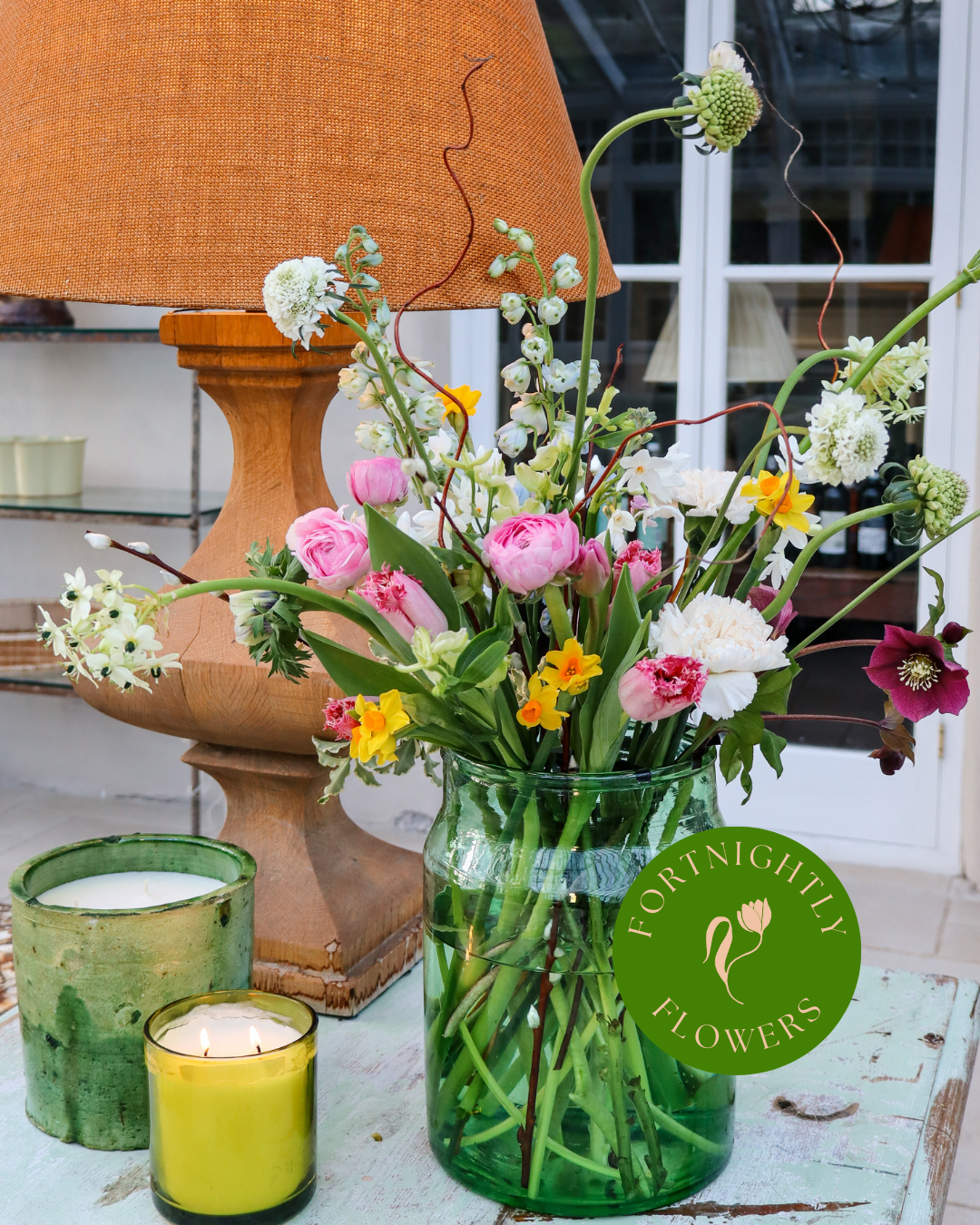 An example of our fortnightly flower service, showcasing our pastel blooms in a green vase, sitting alongside one of our pre lit candles.
