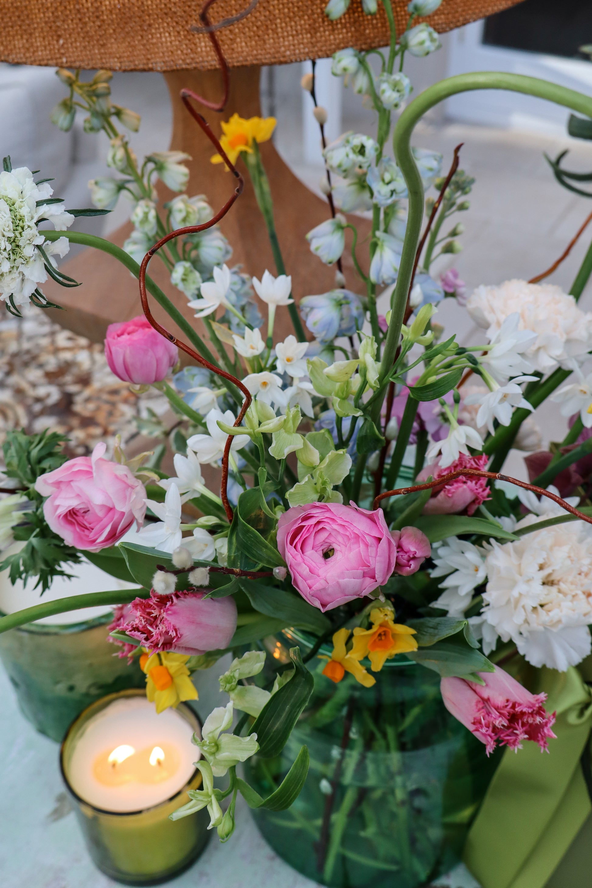 A green glass vase with tulips and daffodils, next to one of our pre-lit candles, showing an example of our luxury bouquets.