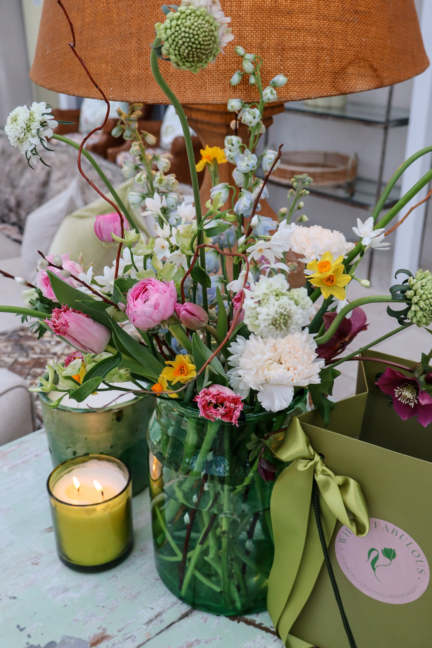 Vibrant spring bouquet in a green vase with pink tulips, white carnations and yellow blooms styled on a pastel table with a green ribboned gift bag and a lit ce from our collection